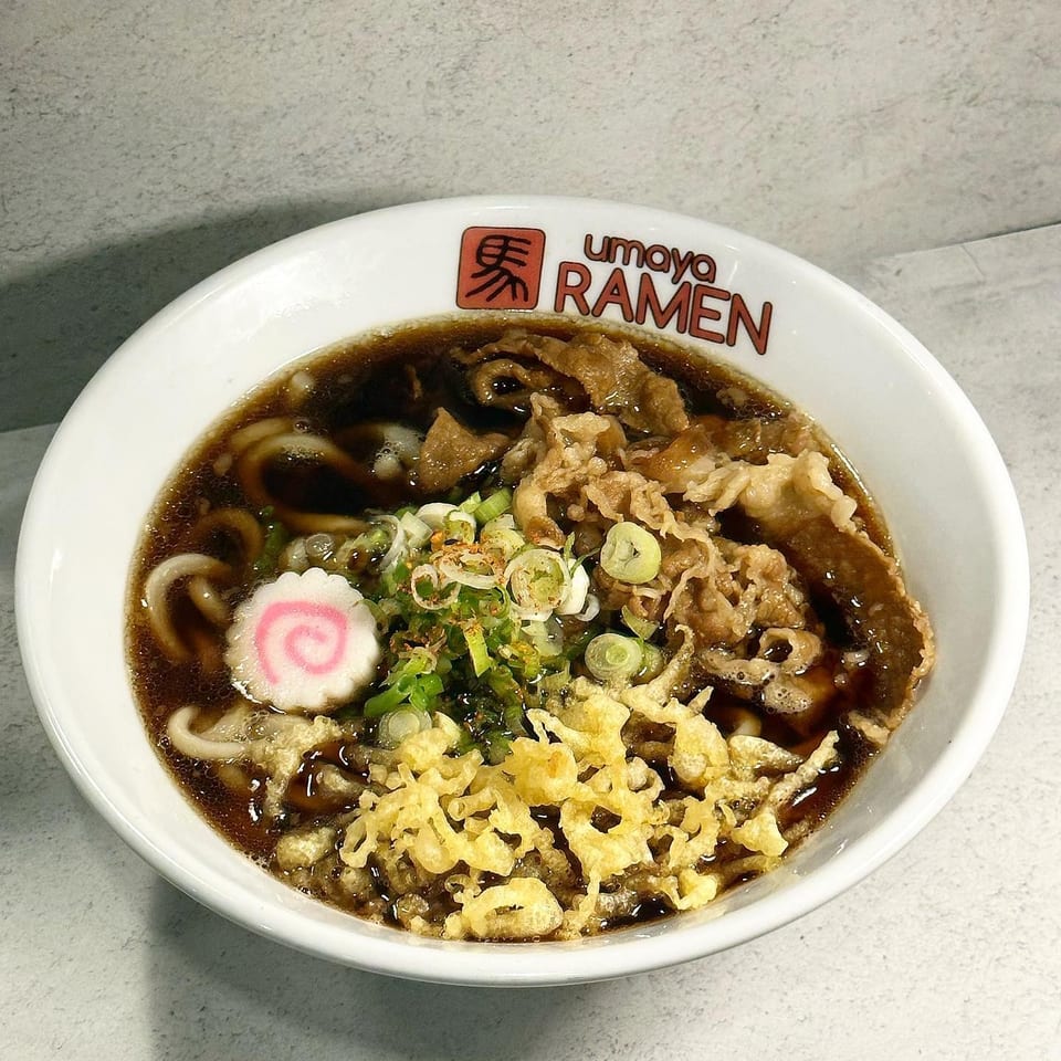 Umaya Ramen bowl with dark broth, noodles, sliced meat, green onions, tempura flakes, and a fish cake.