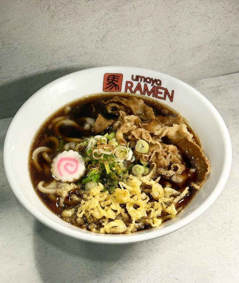 Umaya Ramen bowl with dark broth, noodles, sliced meat, green onions, tempura flakes, and a fish cake.
