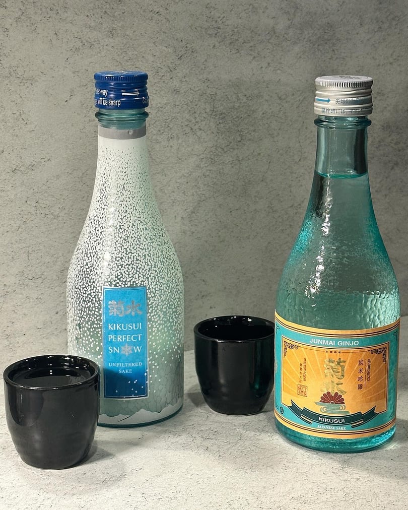 Two bottles of Kikusui sake, one unfiltered and one Junmai Ginjo, with two black sake cups.