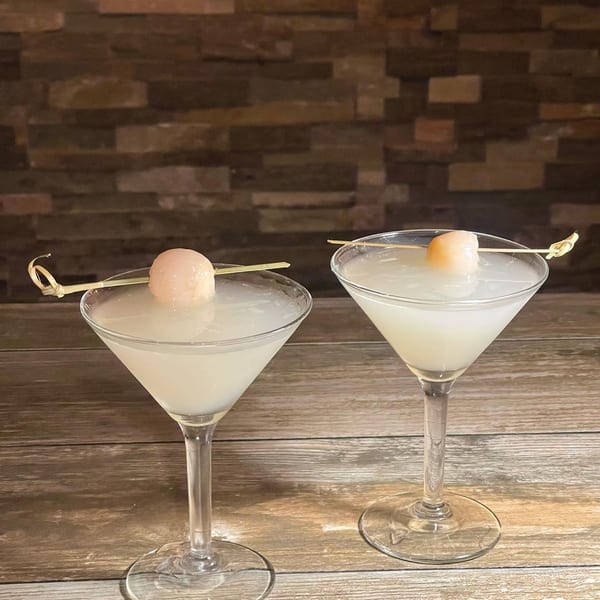 Two lychee martinis garnished with a single lychee on a skewer, against a wood and brick background.