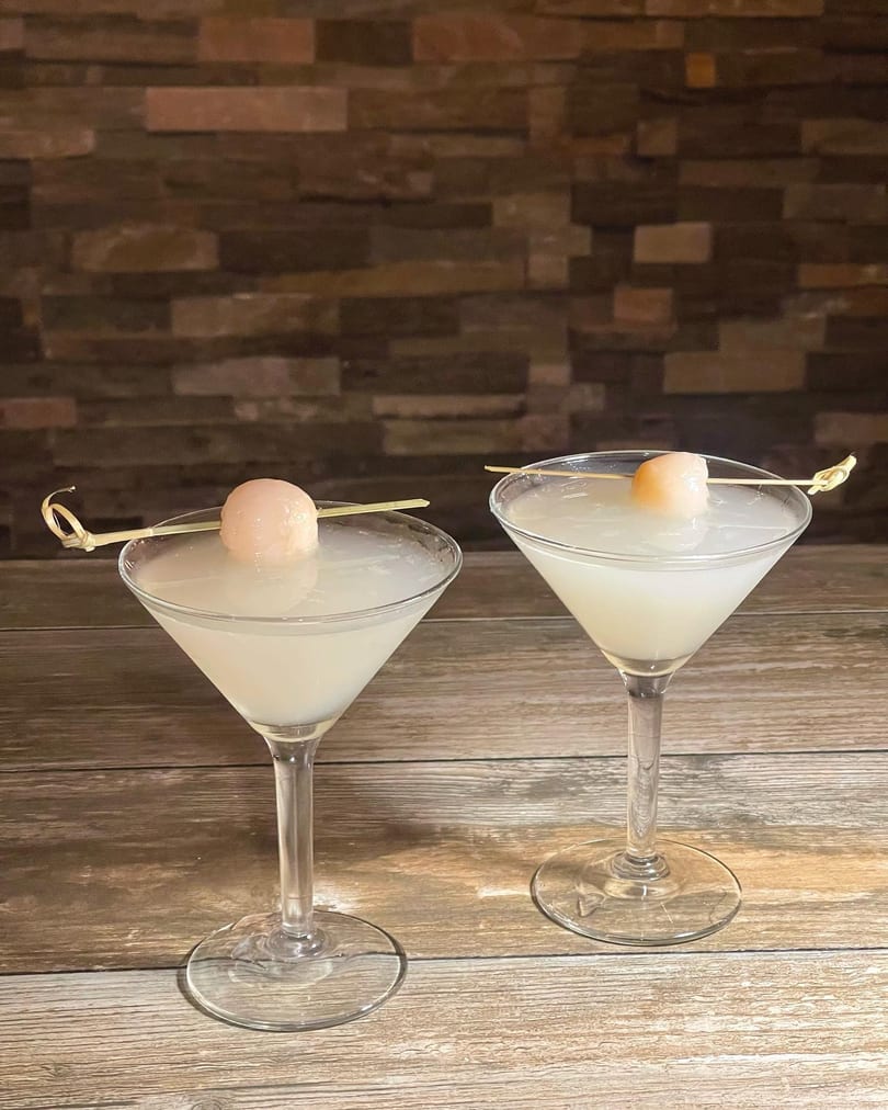 Two lychee martinis garnished with a single lychee on a skewer, against a wood and brick background.