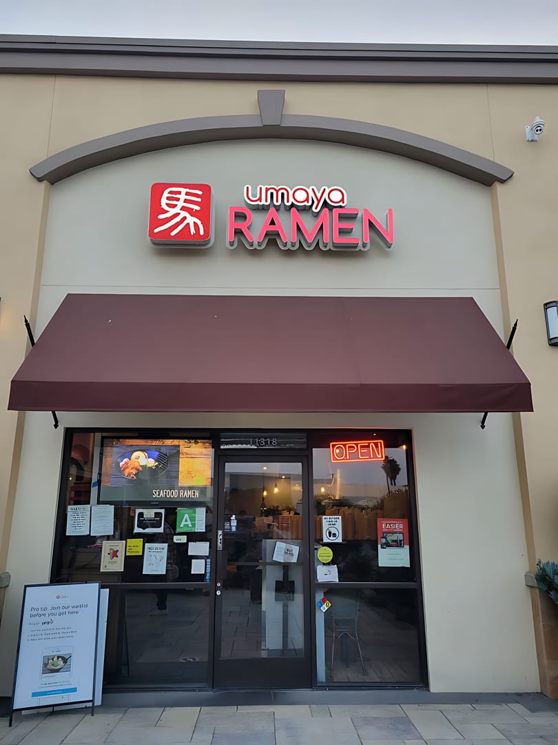 The storefront of Umaya Ramen, featuring a brown awning, neon signs, and an "OPEN" sign.
