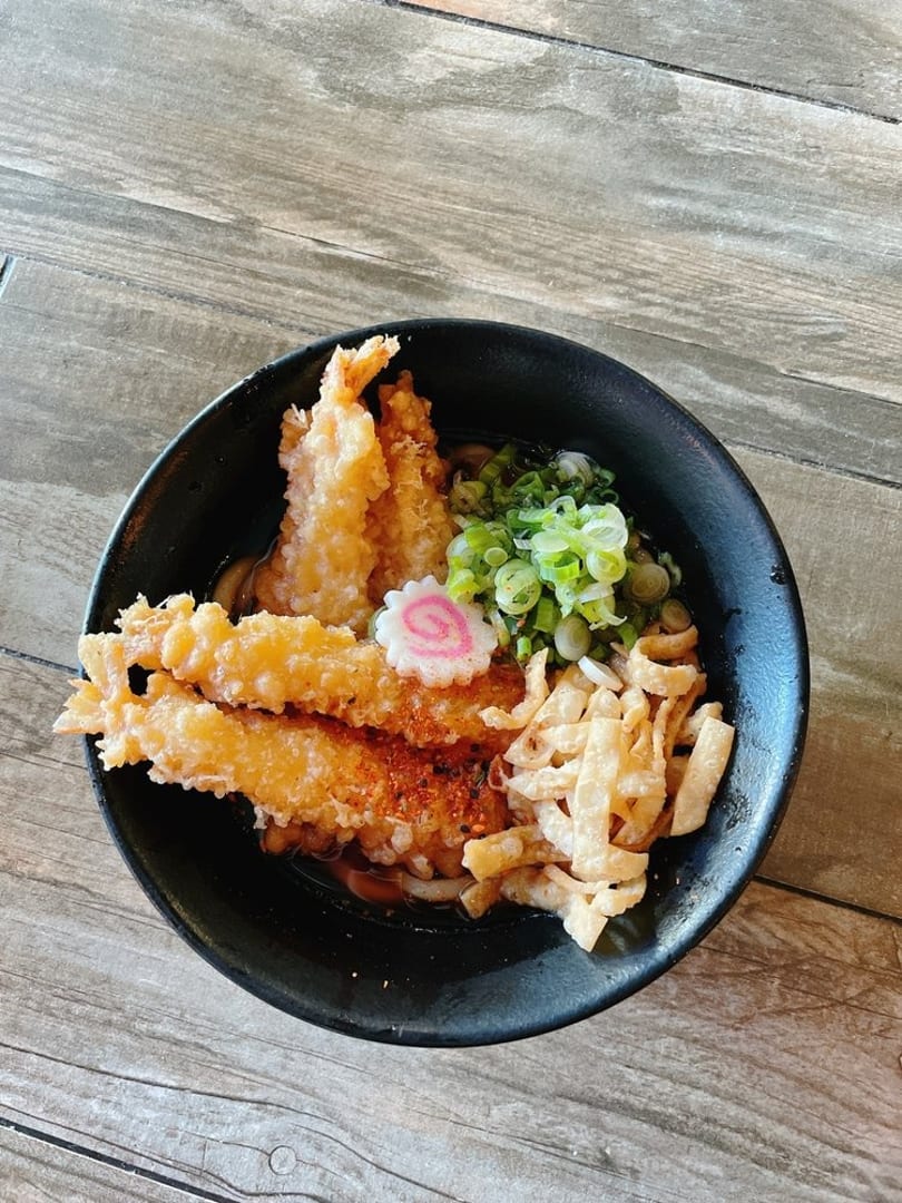 Tempura shrimp and udon noodles with green onions and a pink fish cake in a black bowl.