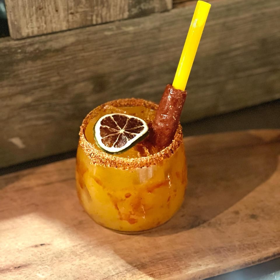 A vibrant yellow drink with a chili-lime rim, dried lime slice, and a tamarind candy straw.