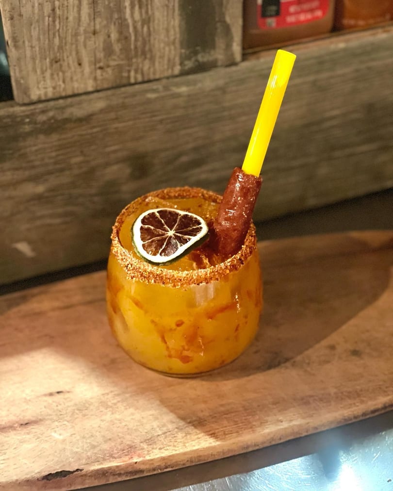A vibrant yellow drink with a chili-lime rim, dried lime slice, and a tamarind candy straw.
