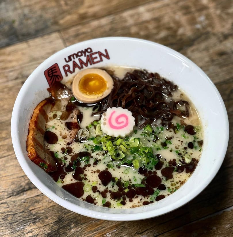 A bowl of Umaya Ramen with pork, egg, mushrooms, green onions, and a pink swirl.