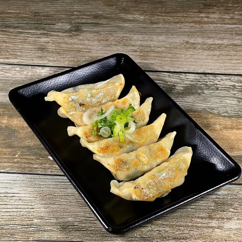 Six golden-brown pan-fried dumplings, garnished with sliced green onions, on a black rectangular plate.