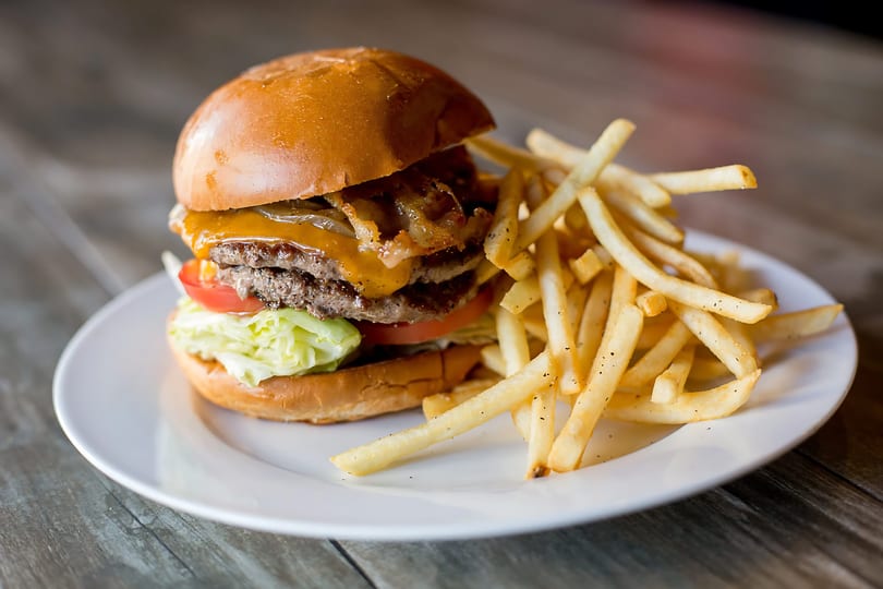 A double cheeseburger with bacon, lettuce, tomato, and fries on a white plate.