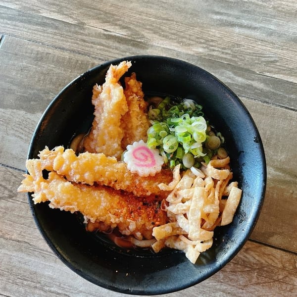 Tempura shrimp and noodles with green onions and a pink swirl fish cake in a black bowl.
