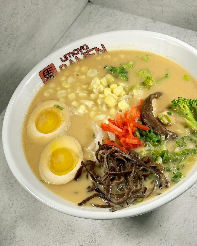 A bowl of Umaya Ramen with broth, corn, broccoli, mushrooms, scallions, and two soft-boiled eggs.