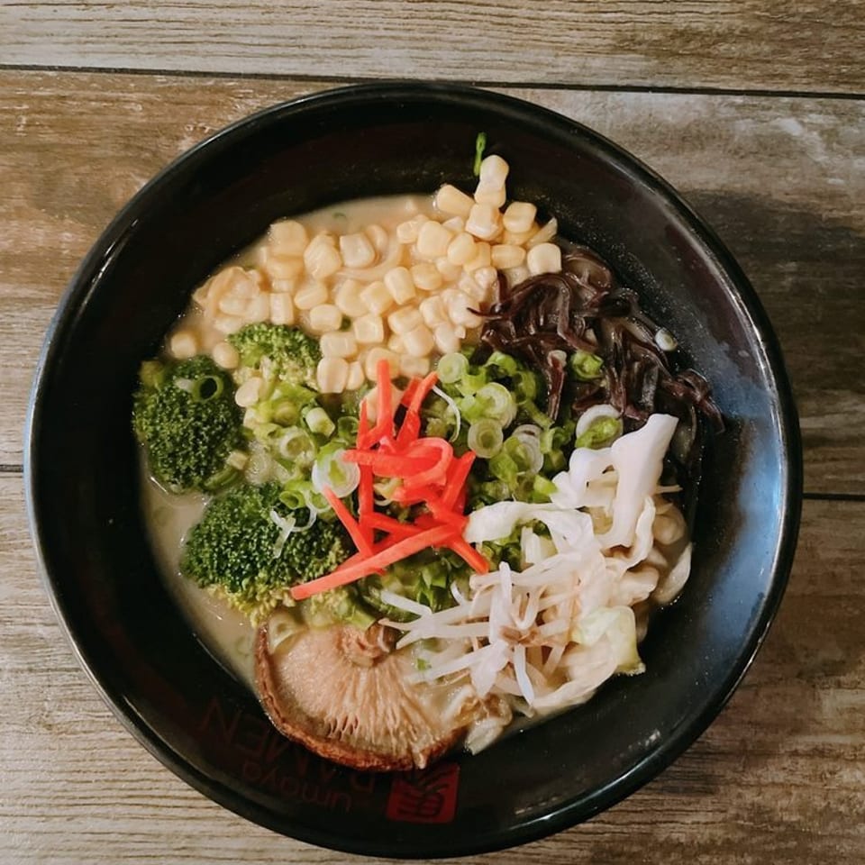 A bowl of ramen with corn, broccoli, mushrooms, and green onions on a wooden table.