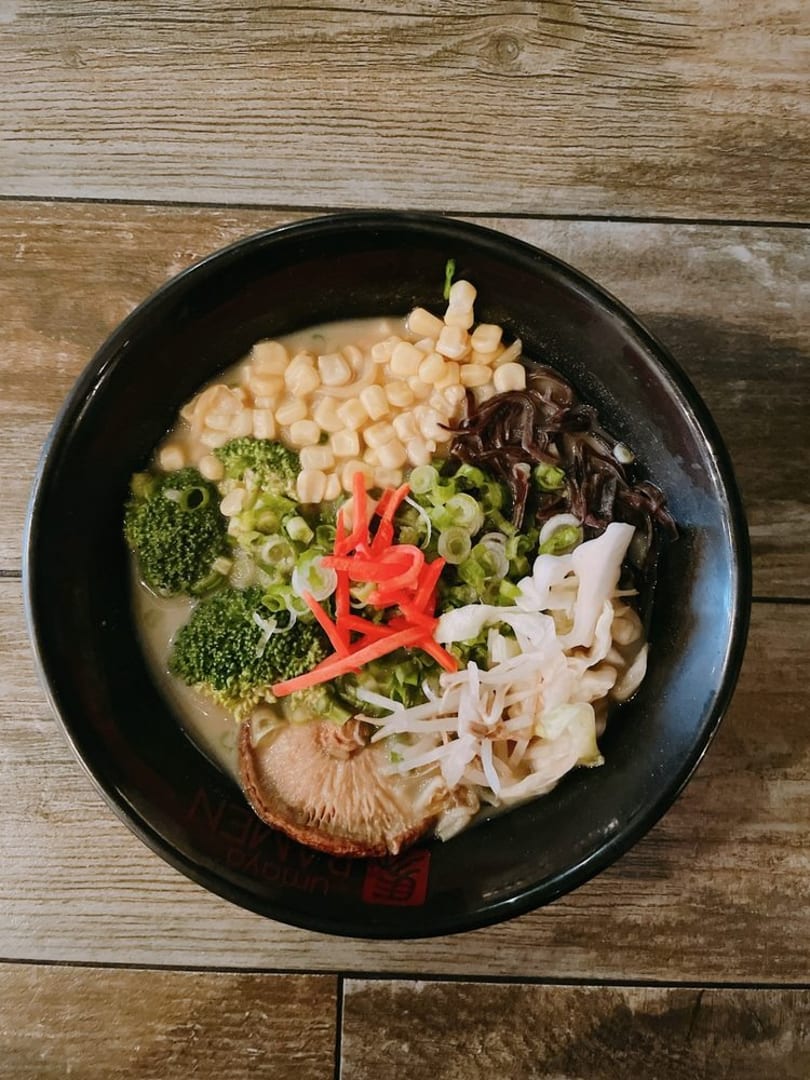 A bowl of ramen with corn, broccoli, mushrooms, and green onions on a wooden table.