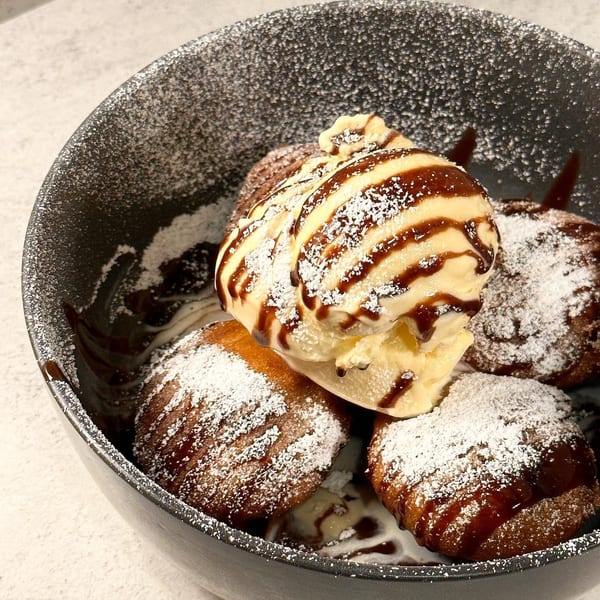 A dark bowl filled with golden-brown donuts, topped with vanilla ice cream, chocolate sauce, and powdered sugar.