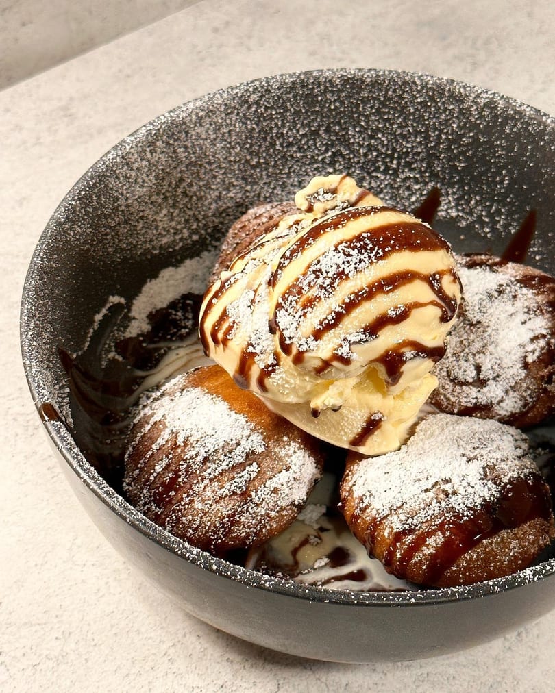 A dark bowl filled with golden-brown donuts, topped with vanilla ice cream, chocolate sauce, and powdered sugar.