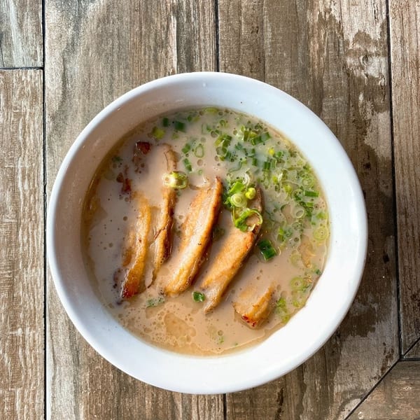 A bowl of ramen with sliced pork belly and green onions in a creamy broth on a wooden table.