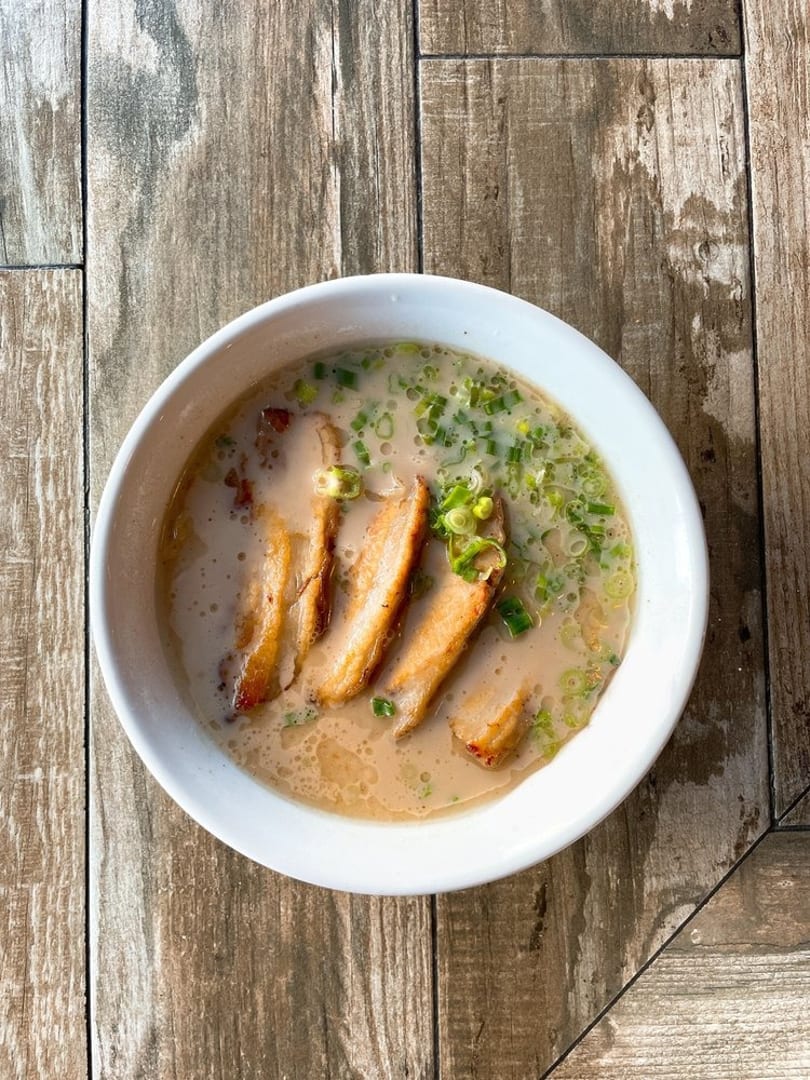 A bowl of ramen with sliced pork belly and green onions in a creamy broth on a wooden table.