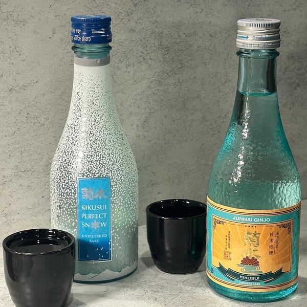 Two bottles of Kikusui sake, one Junmai Ginjo and one Perfect Snow, with two black sake cups.