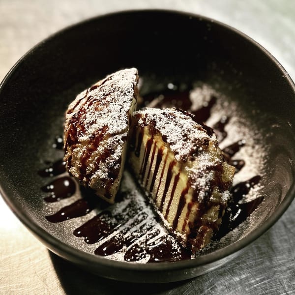 Fried ice cream dessert, cut in half, drizzled with chocolate sauce and powdered sugar in a black bowl.