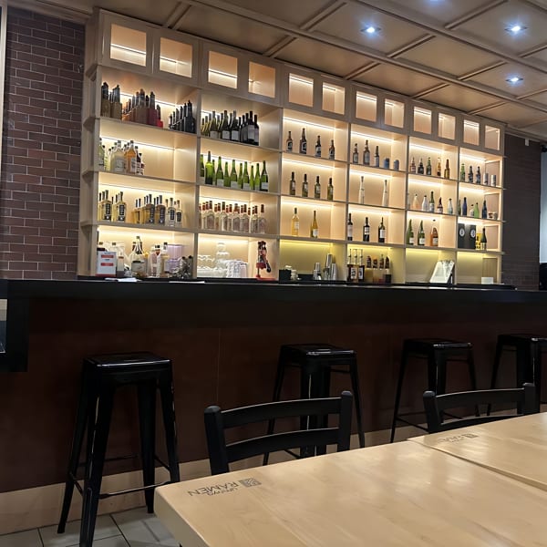 A modern bar area with a large, illuminated shelf displaying numerous bottles, black stools, and light wooden tables.