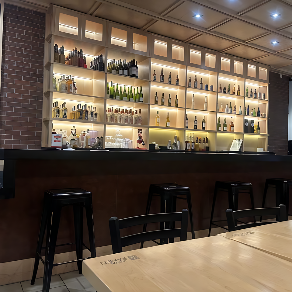 A modern bar area with a large, illuminated shelf displaying numerous bottles, black stools, and light wooden tables.