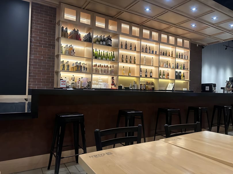 A modern bar area with a large, illuminated shelf displaying numerous bottles, black stools, and light wooden tables.