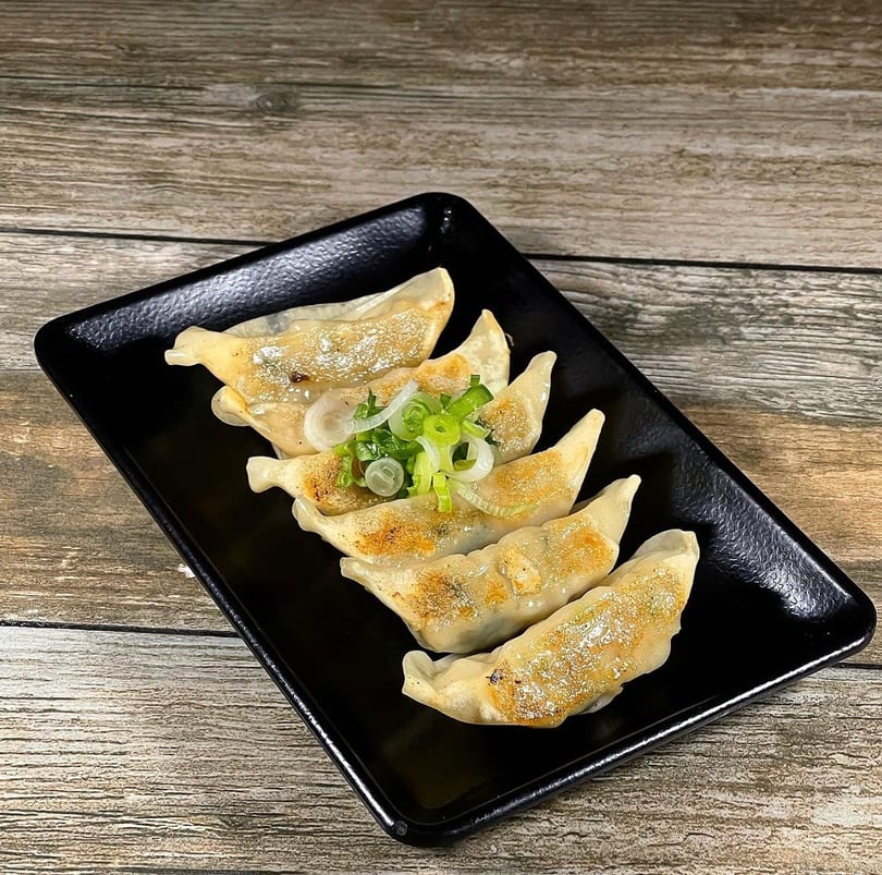 Six pan-fried dumplings with green onions and white onion slices on a black rectangular plate.